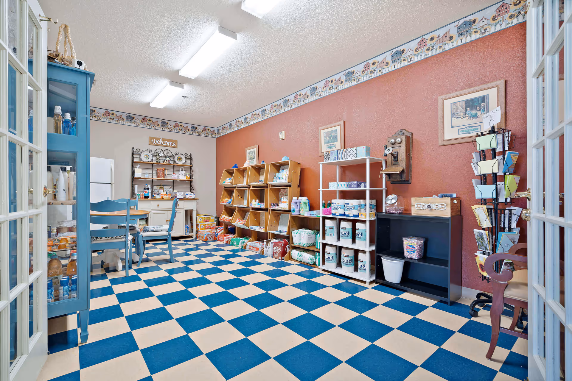 Interior view of a small shop or pantry area with checkered blue and white flooring, blue chairs around a round table, shelves stocked with various snacks, drinks, and household items, a blue cabinet with bottled drinks, and decorative wall art including a vintage telephone and a 'welcome' sign.