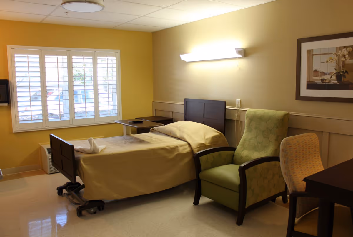 A single bed with beige bedding in a room with yellow walls and a window with white shutters. Next to the bed is a green upholstered armchair and a patterned chair near a dark wooden desk. A framed picture hangs on the wall above the armchair, and a wall-mounted light fixture illuminates the room.