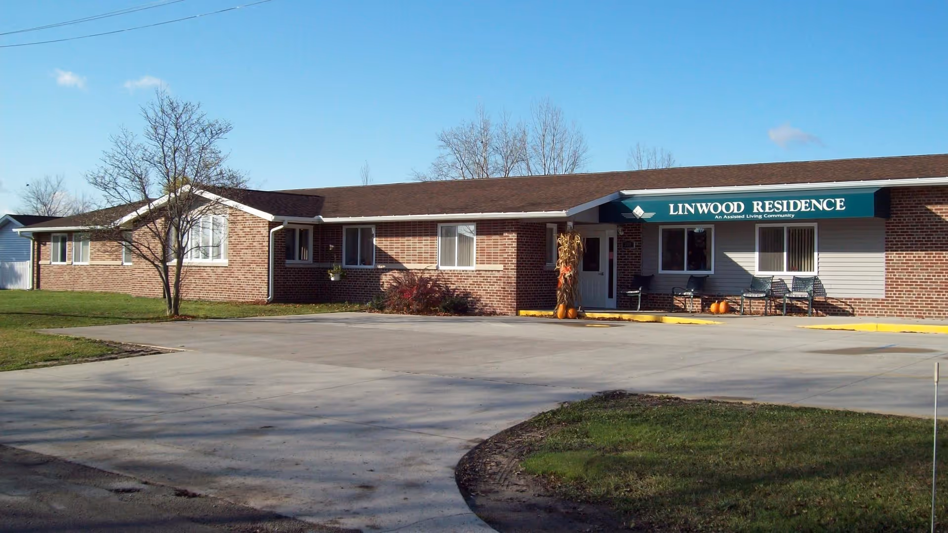 Exterior view of Linwood Residence, a single-story brick assisted living community building with a green awning above the entrance. There are some pumpkins and fall decorations near the door, a few chairs on the porch, and a large paved driveway in front. Trees without leaves are visible in the background under a clear blue sky.