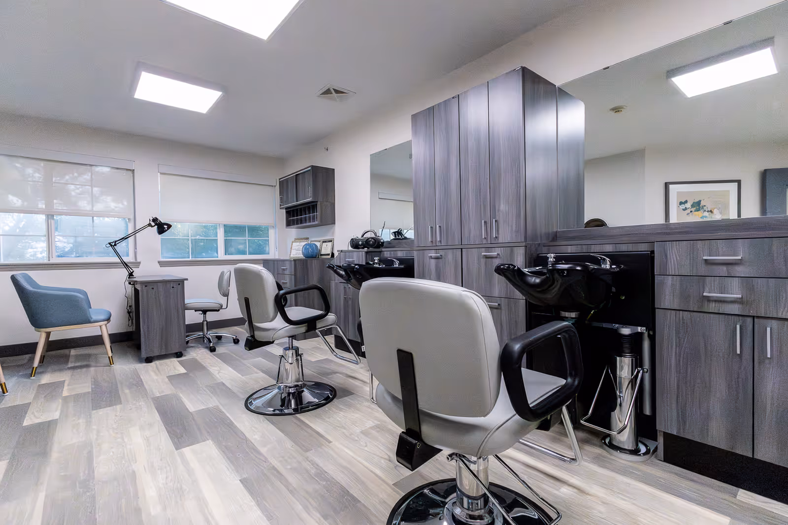 Interior view of a modern salon area in a senior living facility with two salon chairs facing mirrors and washbasins, a desk with a lamp and chair near windows with blinds, and gray wood cabinetry and flooring.