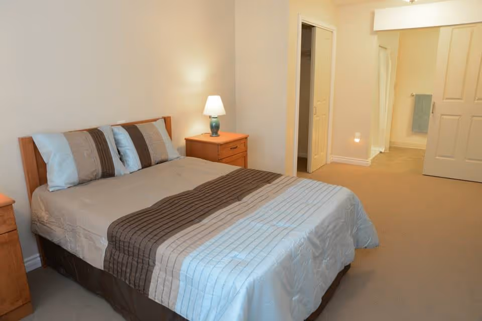 A neatly made bed with striped bedding and three pillows in a bedroom. There are wooden nightstands on either side of the bed, one with a lit table lamp. The room has beige walls and carpet, with an open doorway leading to a hallway and another room with a towel hanging on the wall.