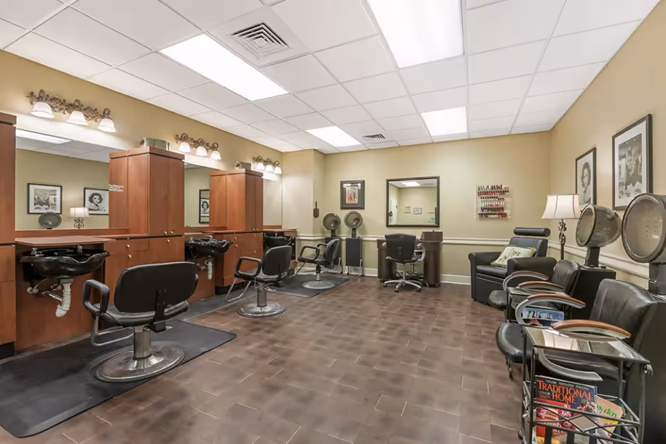 Interior view of a salon area with multiple styling chairs, hair washing sinks, hair dryers, and a nail polish rack on the wall. The room has a tiled floor, beige walls, and framed black and white photos. There are also magazines on a rack near the seating area.