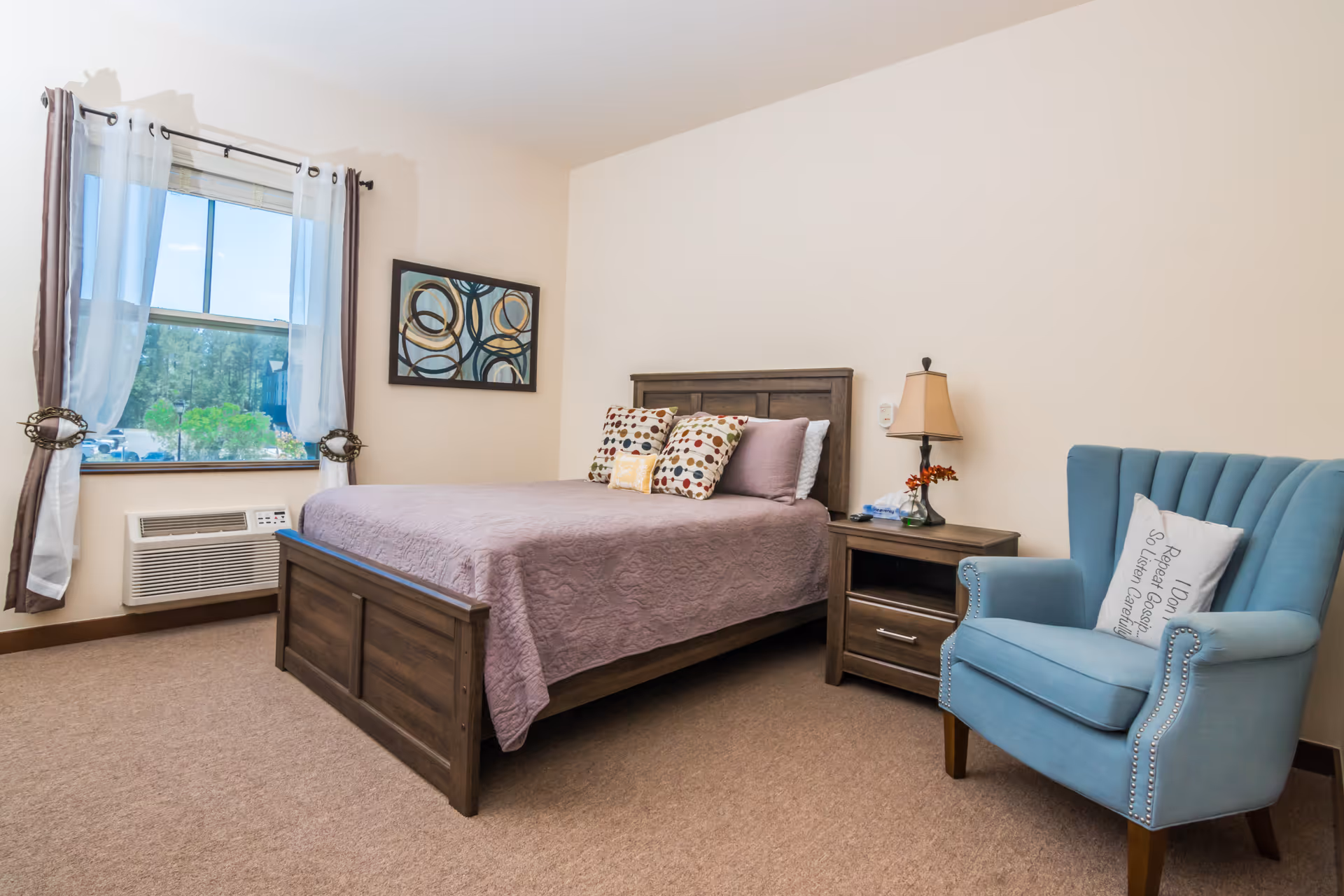 A cozy bedroom in a senior living facility featuring a wooden bed with a lavender quilt and decorative pillows, a wooden nightstand with a lamp and small flower arrangement, a blue upholstered armchair with a decorative pillow, a window with sheer white and brown curtains, and a framed abstract painting on the wall.