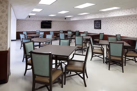 Spacious dining room with multiple square tables and green-upholstered chairs, a wall-mounted TV, and patterned wallpaper.