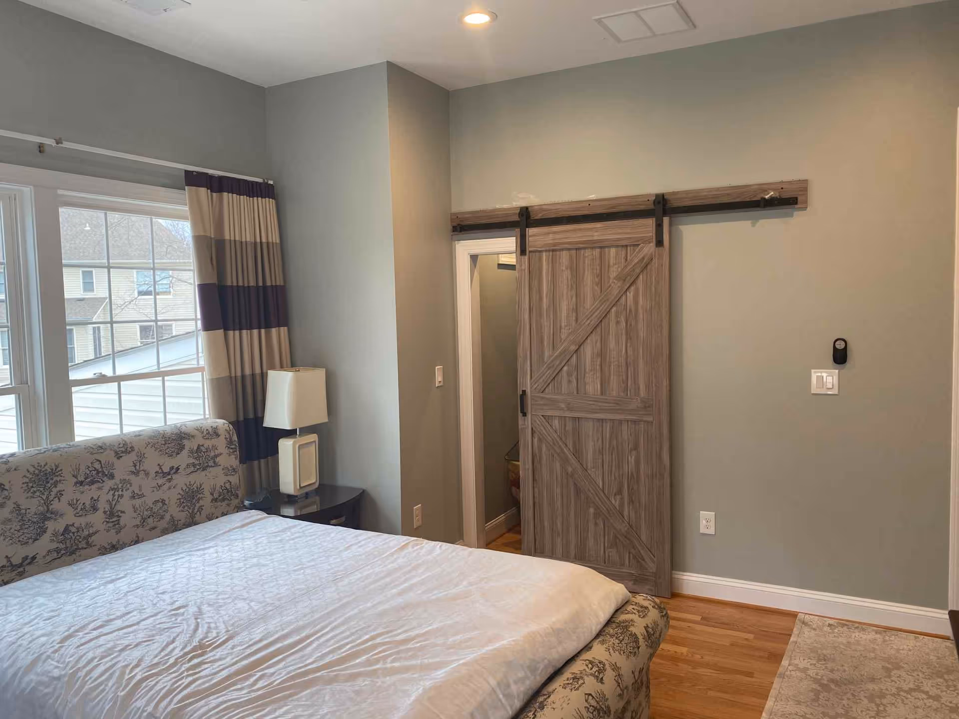 A bedroom with a large window covered by striped curtains, a bed with a patterned headboard and white bedding, a nightstand with a modern lamp, and a sliding barn-style wooden door partially open revealing a small adjoining room.
