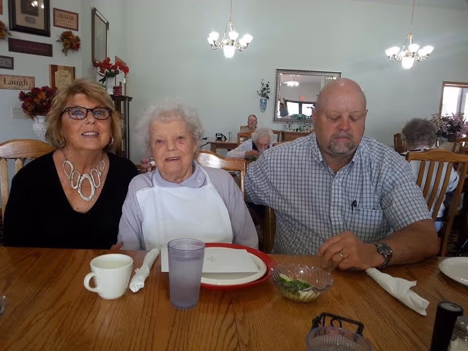Three people sitting at a wooden dining table in a well-lit room. An elderly woman wearing a white bib sits in the center, flanked by a woman with glasses and a necklace on the left and a man in a checkered shirt on the right. The table has a white cup, a glass of water, a plate, and a small bowl with salad. In the background, other elderly individuals are seated at tables, with wall decorations and chandeliers visible.