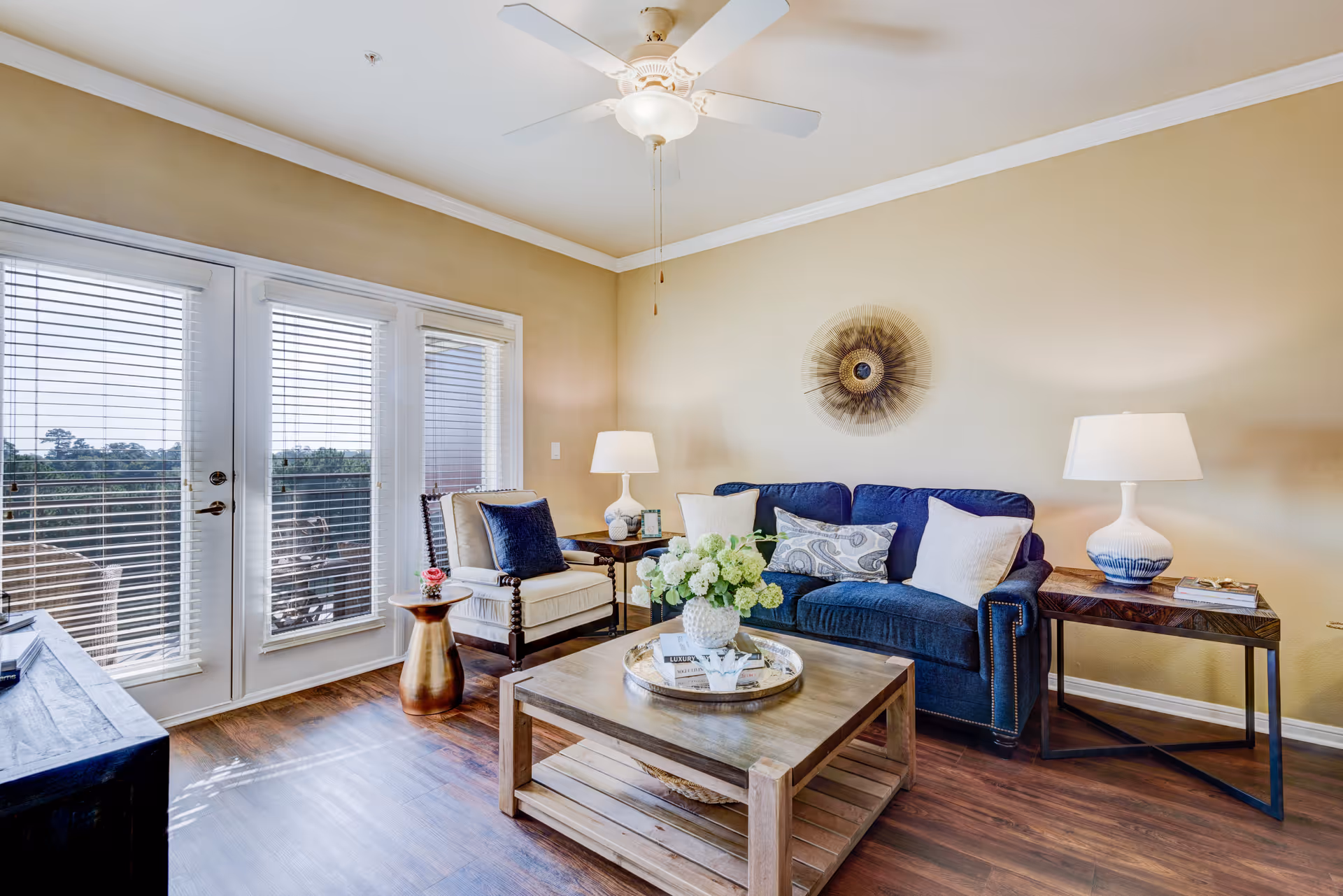A bright living room with a blue sofa adorned with multiple cushions, a wooden coffee table with a flower vase and books, a beige armchair with a dark blue cushion, two side tables each with a lamp, and large glass doors with blinds leading to an outdoor balcony.