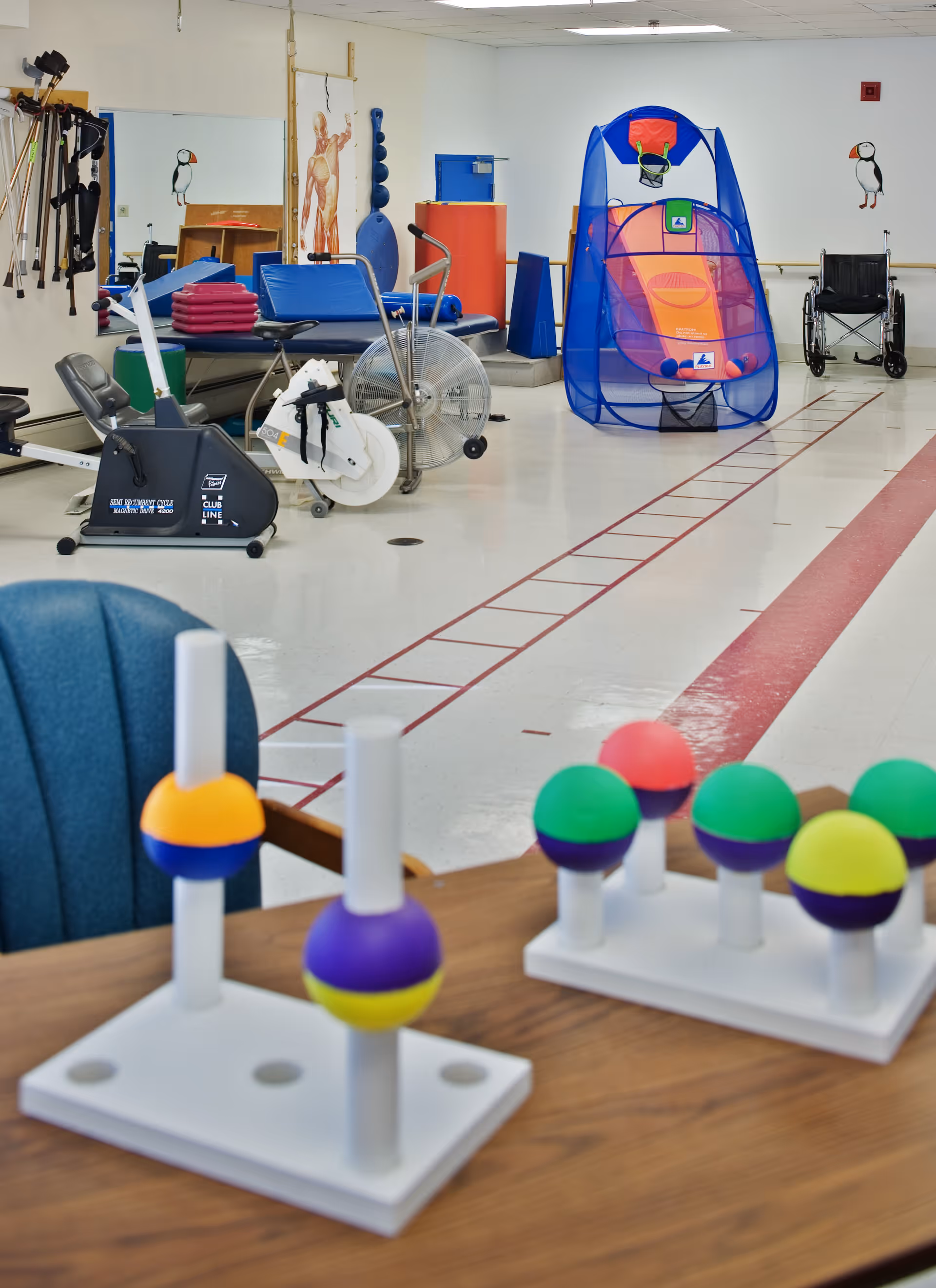 A physical therapy or rehabilitation room with exercise equipment including a stationary bike, therapy tables, and a basketball shooting game. In the foreground, there are colorful pegboards with balls on a wooden table. The room has white walls with anatomical posters and a wheelchair is visible in the background.