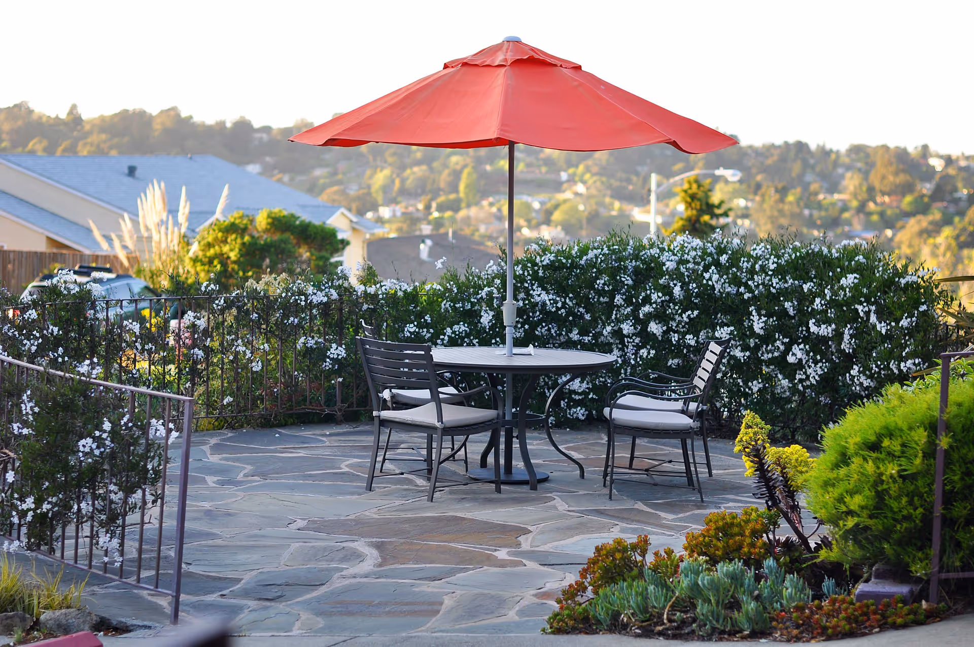 Patio with a round table, four chairs and a red umbrella surrounded by flowering shrubs and a distant view of houses.