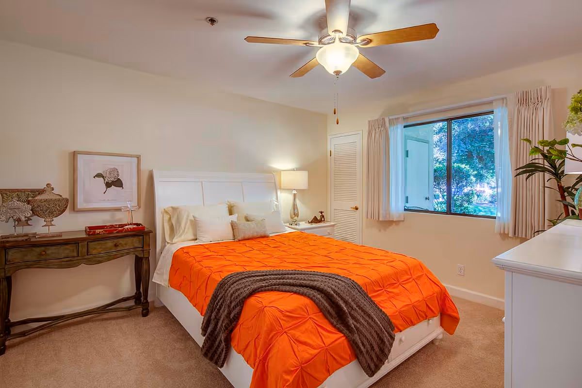 A cozy bedroom with a white bed frame and an orange bedspread. There are multiple pillows on the bed and a dark gray throw blanket at the foot. A wooden side table with decorative items and a framed flower picture is on the left side. A white nightstand with a lamp and small decor is on the right side. A window with beige curtains shows greenery outside. The ceiling has a fan with a light fixture.