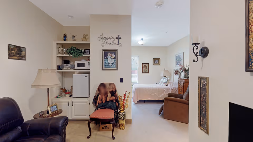 Interior view of a senior living facility room showing a cozy living area with a leather armchair, a side table with a lamp and framed photo, a chair with a blanket, and built-in shelves holding a microwave, mini fridge, and decorative items. In the background, there is a bedroom with a bed, nightstand with lamp, dresser, and wall decorations.