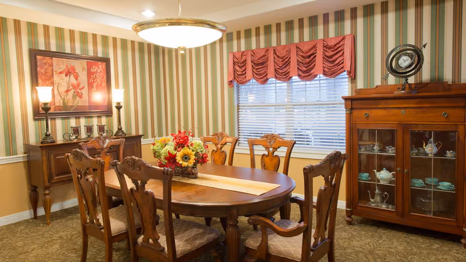 A traditional dining room with a wooden oval dining table surrounded by six carved wooden chairs with cushioned seats. The table has a floral centerpiece and a beige runner. The room features striped wallpaper in green, beige, and white tones, a window with white blinds and a red valance, a wooden sideboard with two lamps and a framed floral painting above it, and a wooden cabinet displaying a tea set and a decorative clock on top.