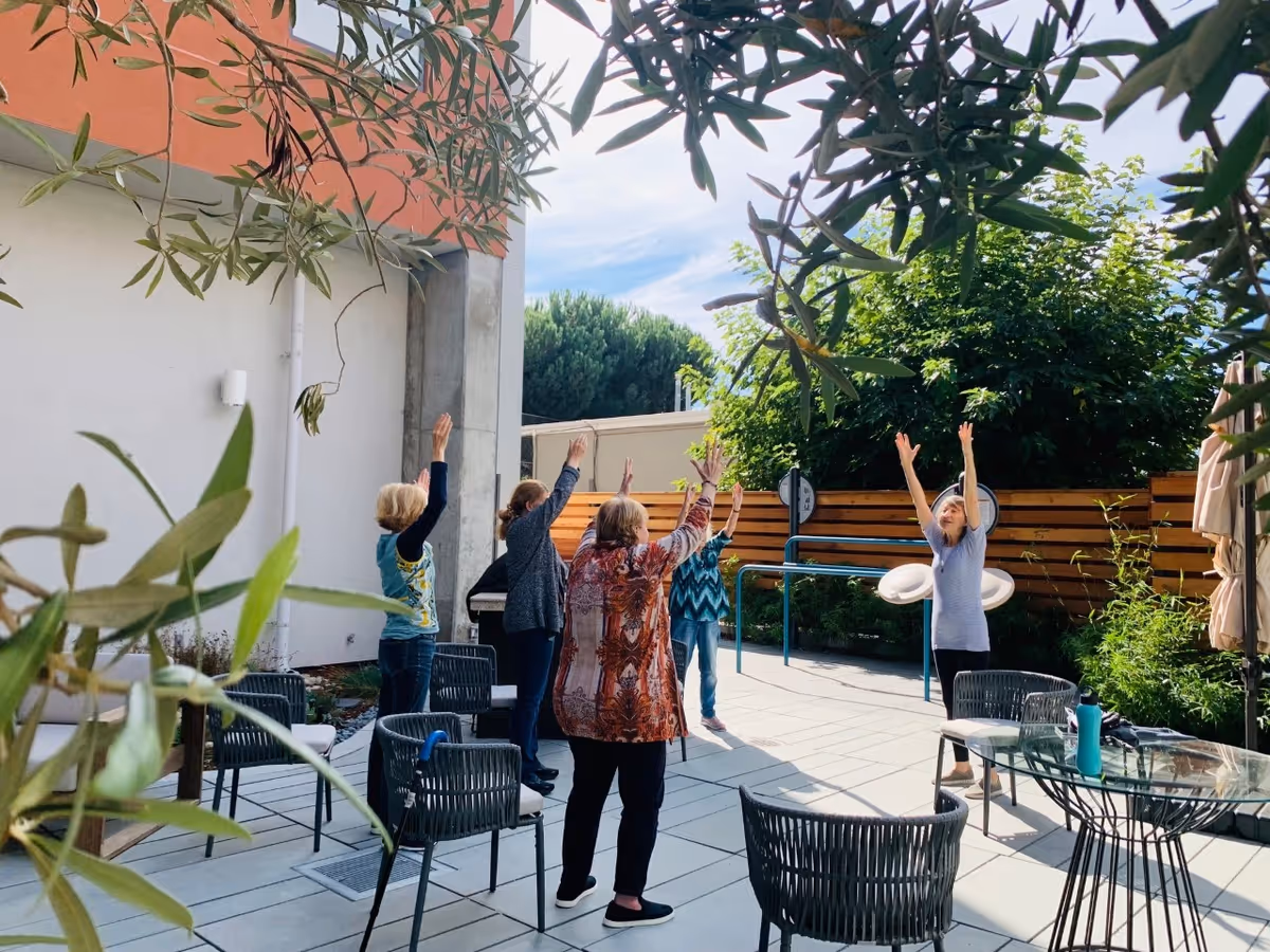 A group of elderly women standing in a circle outdoors on a patio, raising their arms upwards as if participating in a stretching or exercise activity. The patio is surrounded by greenery, a wooden fence, and some outdoor furniture including chairs and a glass table. The sky is partly cloudy and the setting appears peaceful and sunny.