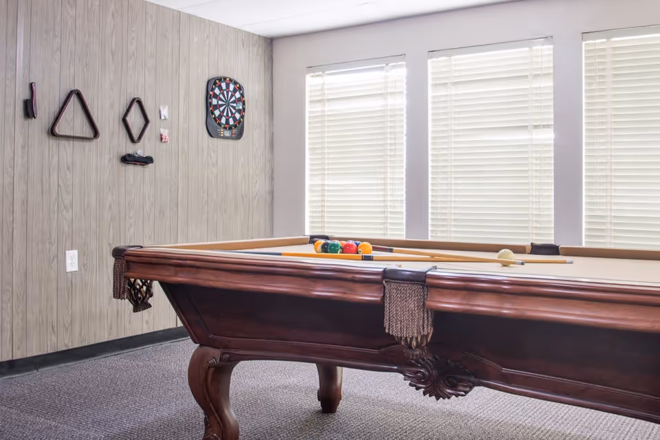 A game room with a wooden pool table featuring billiard balls and cues on top. The room has a wall-mounted dartboard and racks holding pool cues and chalk. Three large windows with white blinds allow natural light to fill the space.