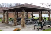 Outdoor covered pavilion with several tables and chairs arranged underneath. The pavilion is decorated with bundles of dried corn stalks tied to the supporting posts. Surrounding the pavilion are benches and a grassy area with trees and buildings in the background.