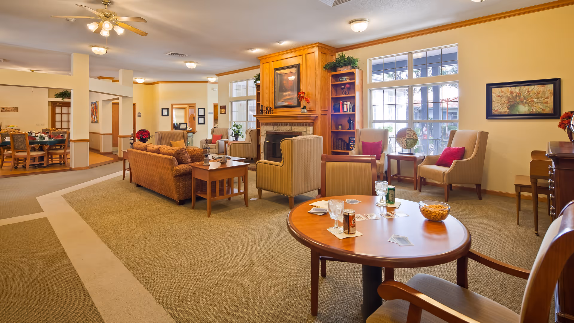 A cozy senior living facility common area with comfortable armchairs and sofas arranged around a wooden coffee table. The room features a fireplace with a wooden mantel, built-in bookshelves, large windows letting in natural light, and a dining area visible in the background. The decor includes framed artwork, plants, and a ceiling fan with lights.