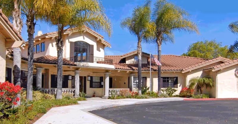 Exterior view of a single-story building with a tiled roof, several palm trees, and landscaped bushes and flowers in front. The building has a covered entrance supported by columns and an American flag on a flagpole near the entrance under a clear blue sky.