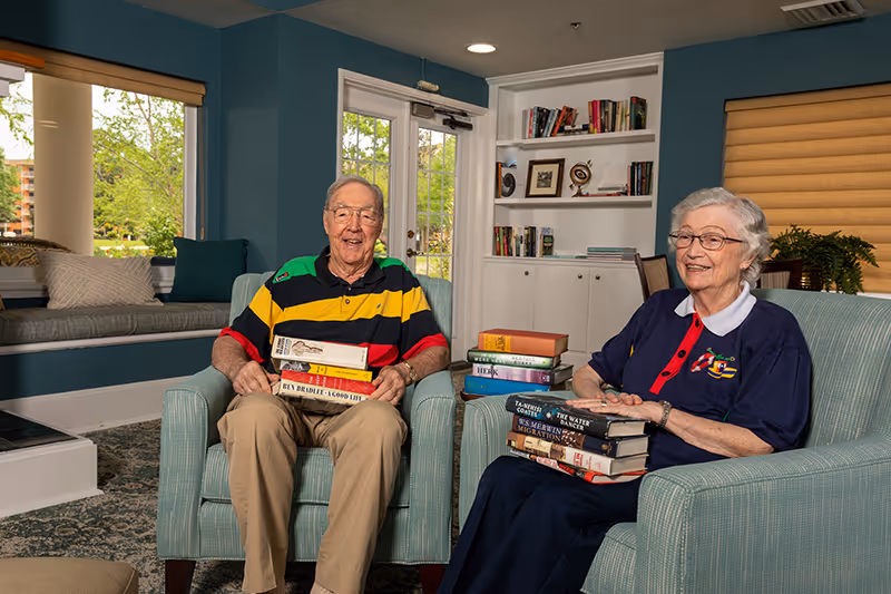 Two elderly individuals sitting in armchairs in a cozy living room area, each holding a stack of books. The room features a built-in bookshelf with books and decorative items, large windows with beige blinds, and a cushioned window seat with pillows. The walls are painted teal, and there is a glass door leading outside.