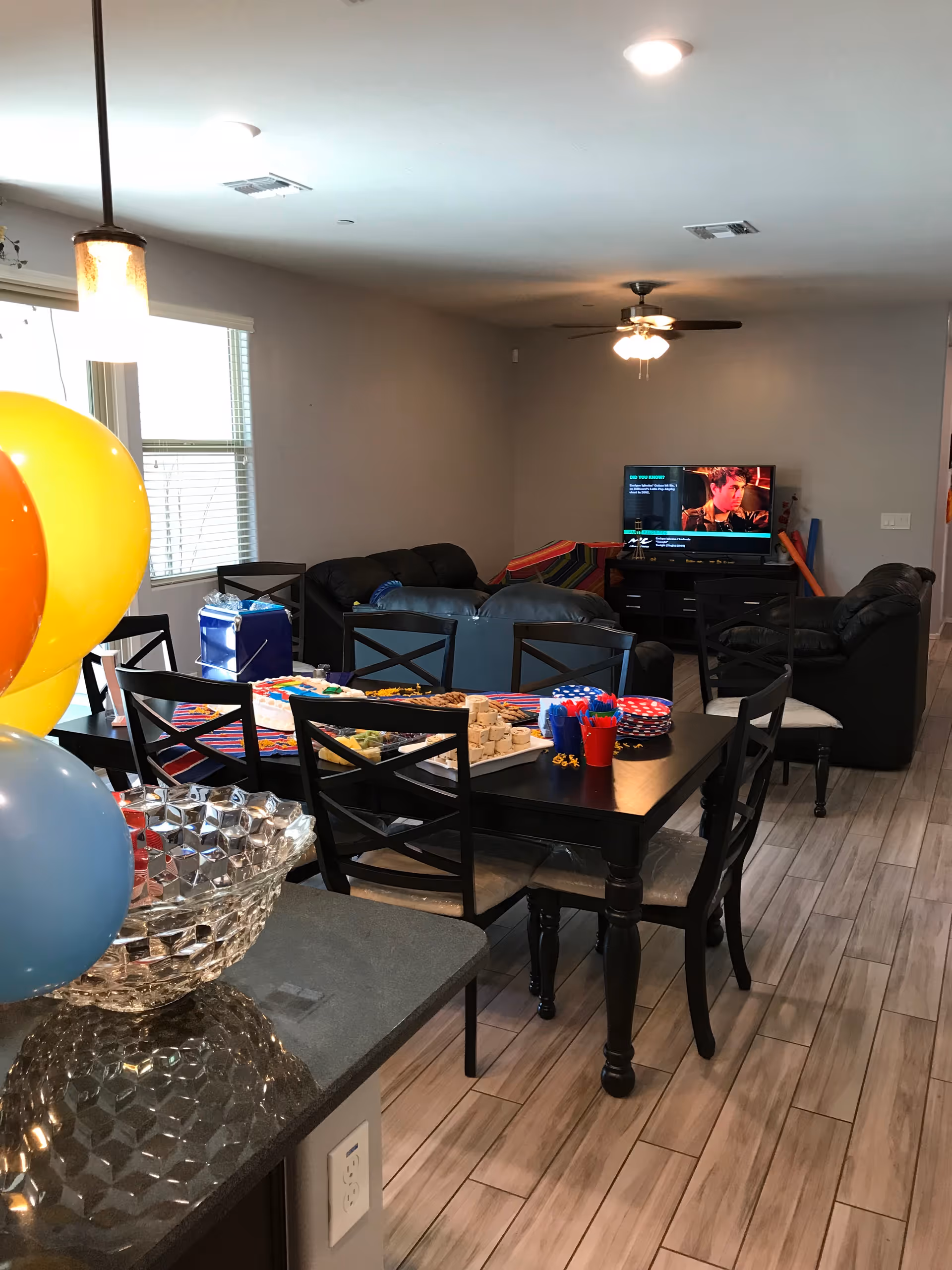 Interior view of a senior living facility common area with a dining table set for a party, including colorful balloons, plates, and snacks. In the background, there are black leather sofas arranged around a TV mounted on a stand, with a ceiling fan overhead and windows letting in natural light.