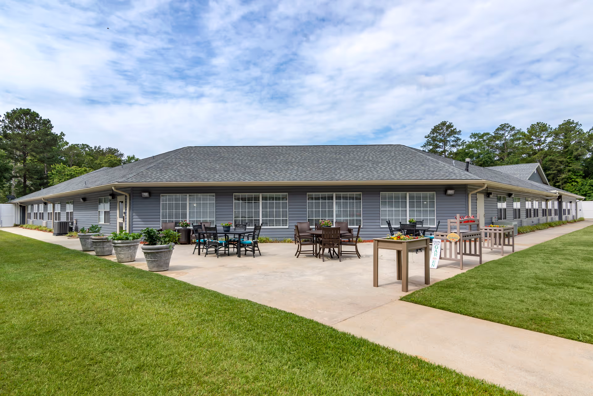 Outdoor patio area of a senior living facility with several tables and chairs arranged on a concrete surface. There are large potted plants and raised garden beds with flowers and greenery. The building has gray siding and multiple windows with white blinds. The sky is partly cloudy and there is green grass surrounding the patio.