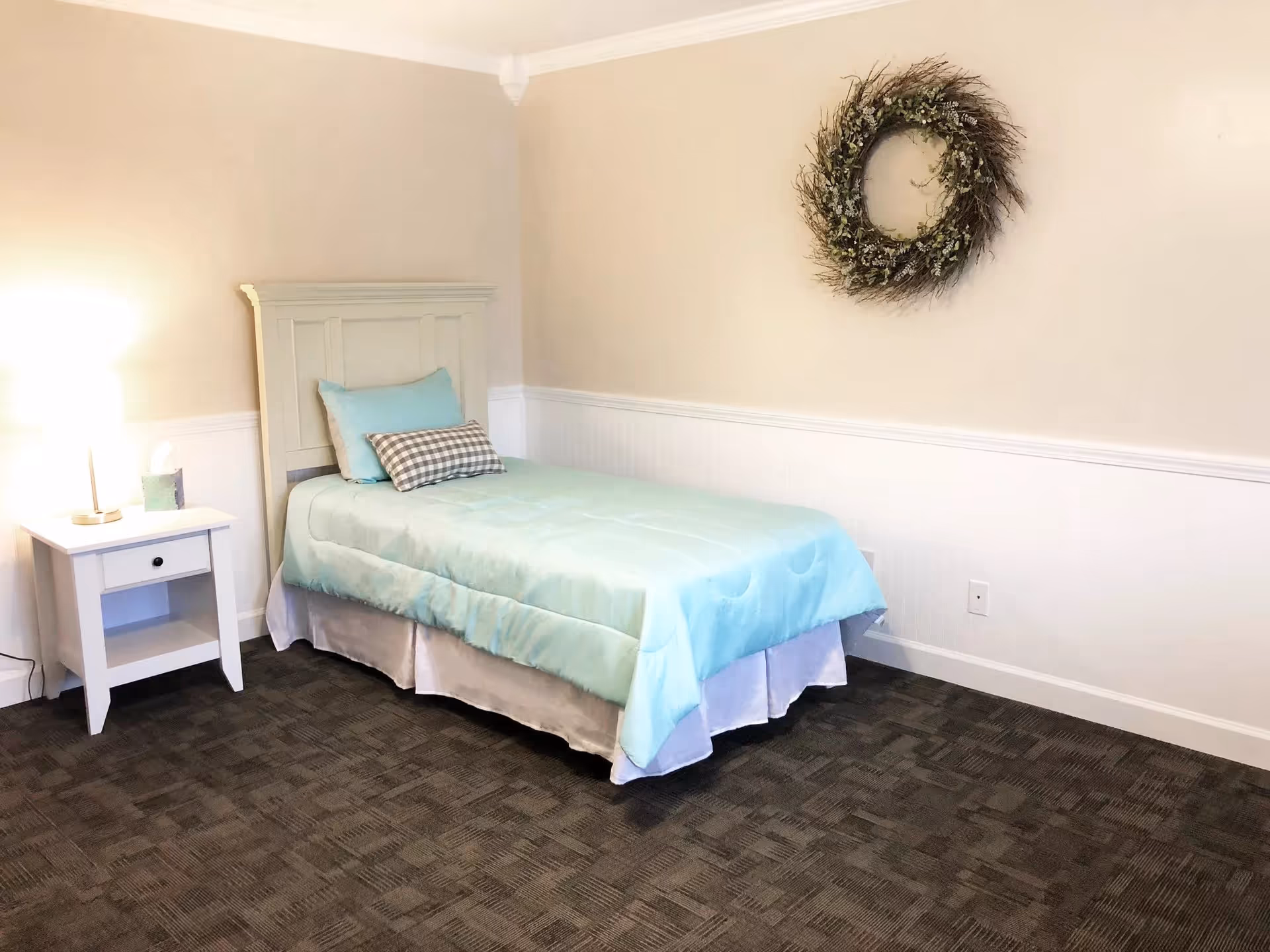 A simple bedroom with a single bed featuring a light blue comforter and two pillows, one solid blue and one checkered. There is a white nightstand with a lamp and a box of tissues on it. The walls are beige with white wainscoting, and a decorative wreath hangs on the wall above the bed. The floor is carpeted in a dark pattern.