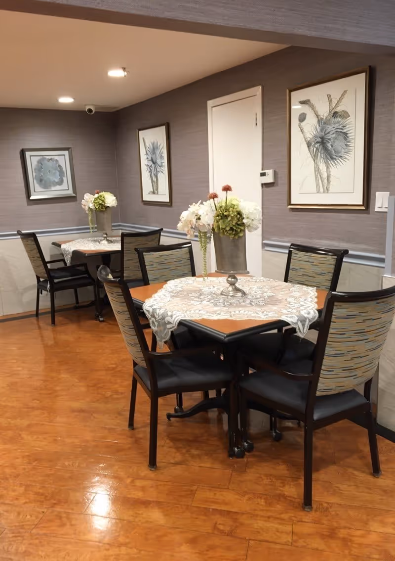 A cozy dining area with two wooden tables covered with lace tablecloths, each surrounded by four cushioned chairs with patterned backs. Each table has a floral centerpiece in a metallic vase. The walls are decorated with framed botanical artwork, and the floor has a polished wood finish.