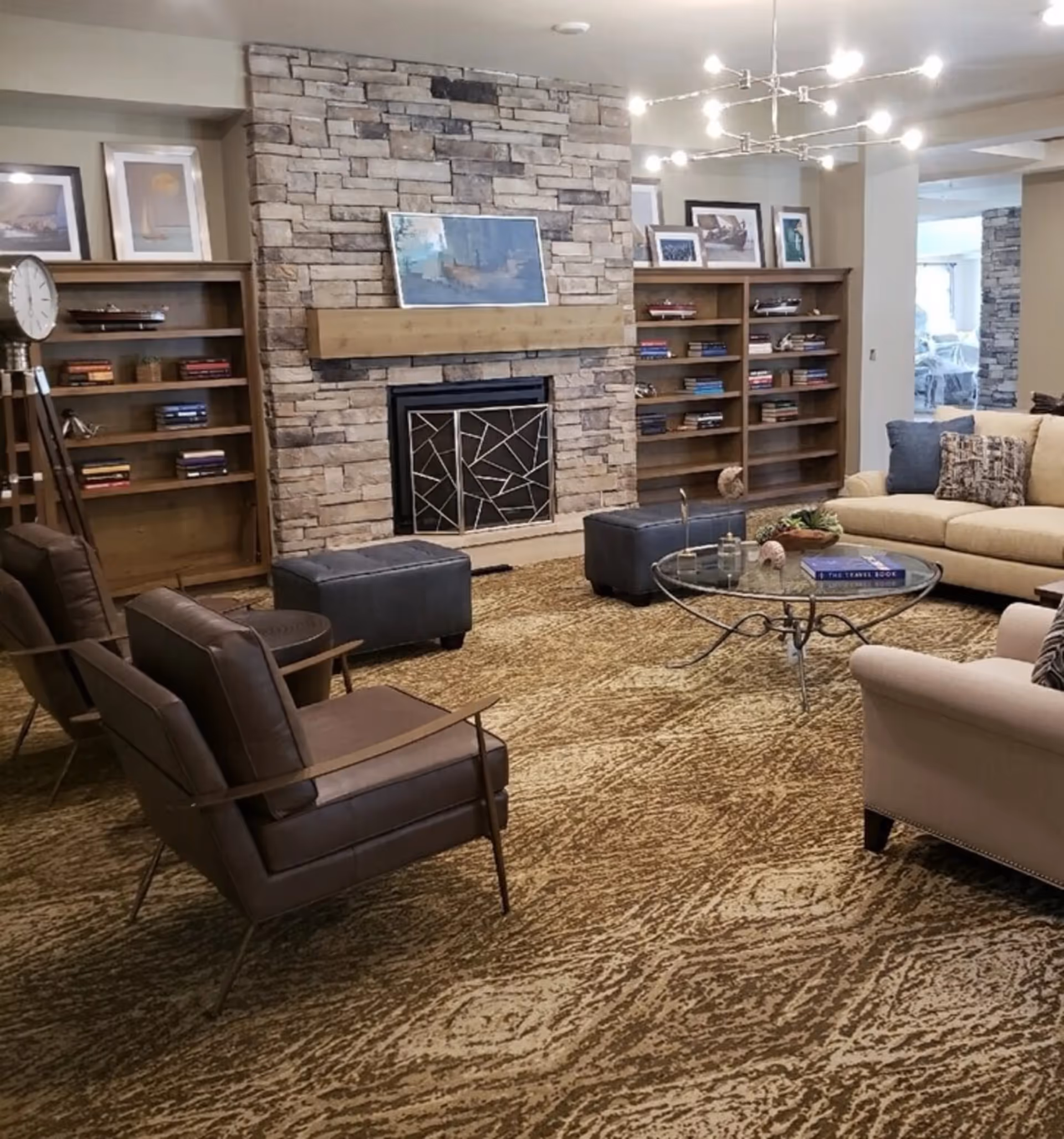 A cozy living room with a stone fireplace in the center, flanked by wooden bookshelves filled with books and decorative items. There are two dark brown leather chairs, two dark ottomans, a glass coffee table with decorative items, and a beige sofa with cushions. The room has a patterned carpet and modern ceiling light fixtures.