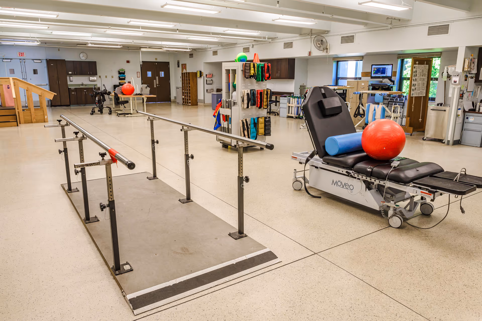 A spacious rehabilitation room with parallel bars for walking exercises, a therapy table with a red exercise ball and blue foam roller on it, various therapy equipment and supplies on shelves, and a set of stairs in the background. The room has bright lighting and a clean, open layout.