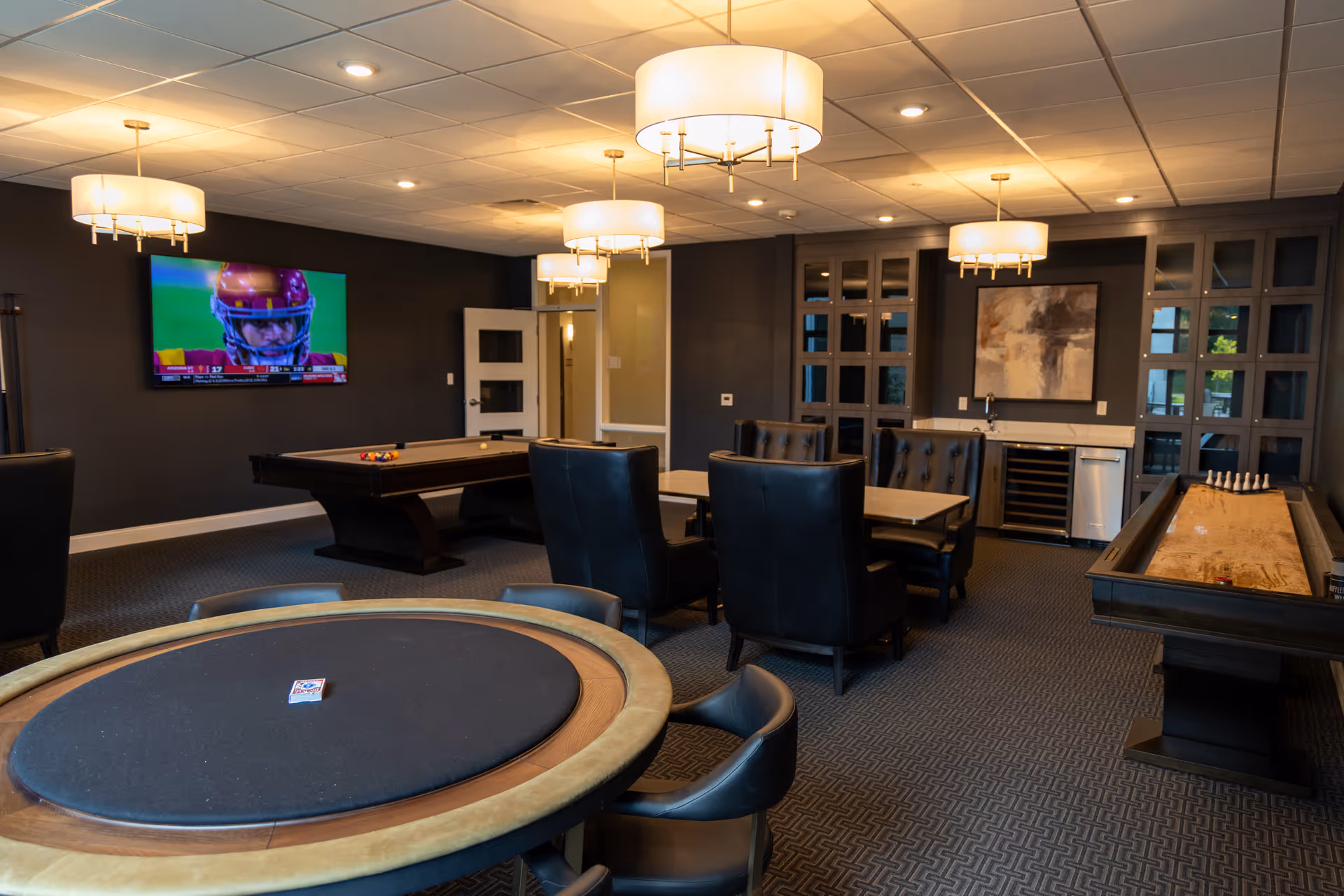 A spacious game room with a poker table in the foreground, a pool table in the middle, and a shuffleboard table on the right. The room features dark walls, carpeted floor, modern ceiling lights, a wall-mounted TV showing a football game, and a small bar area with a wine fridge and cabinets.