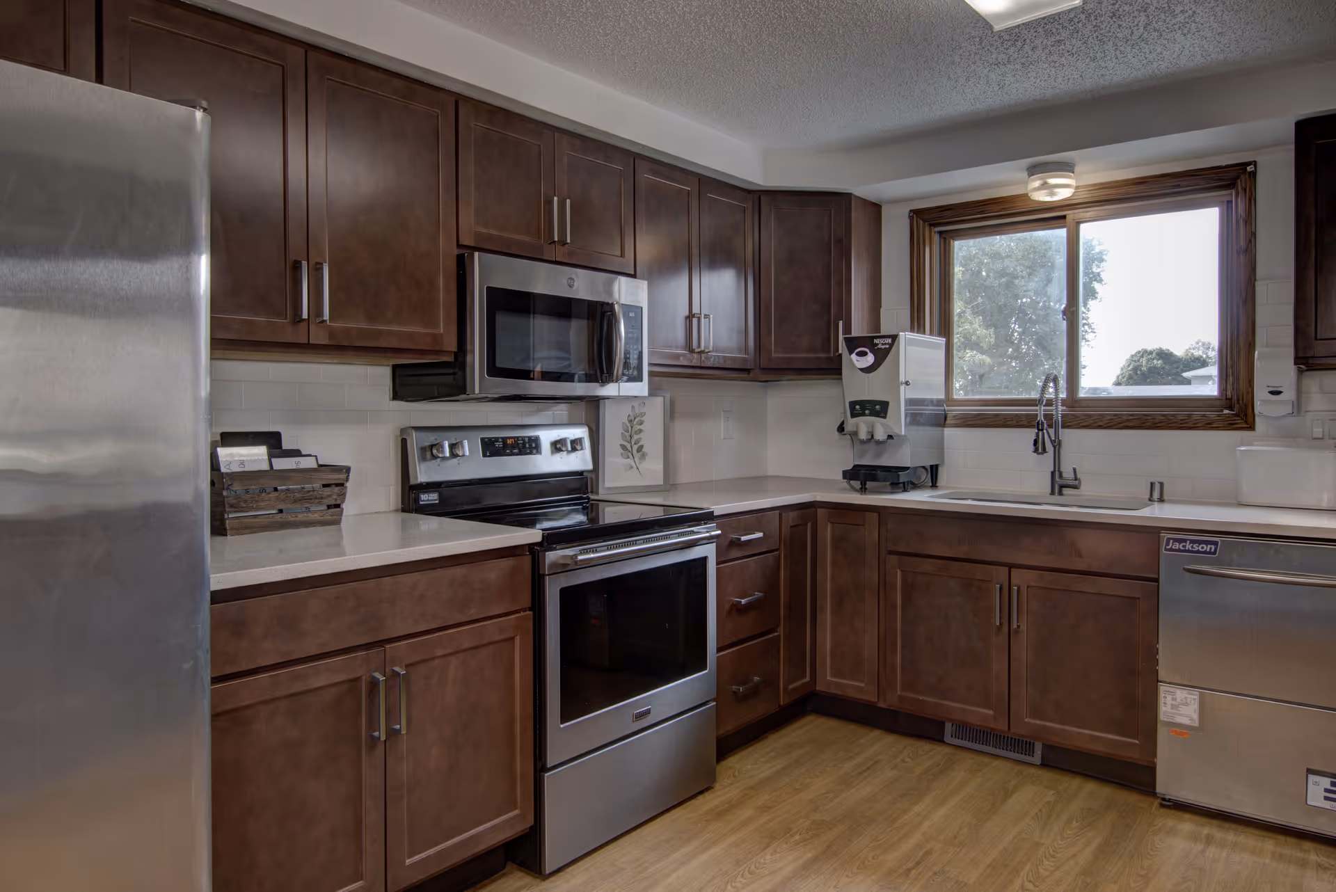 Modern kitchen with stainless steel refrigerator, oven, microwave, and dishwasher. Dark wood cabinets and white countertops surround the appliances. A window above the sink lets in natural light, and a coffee machine is placed on the counter near the window.