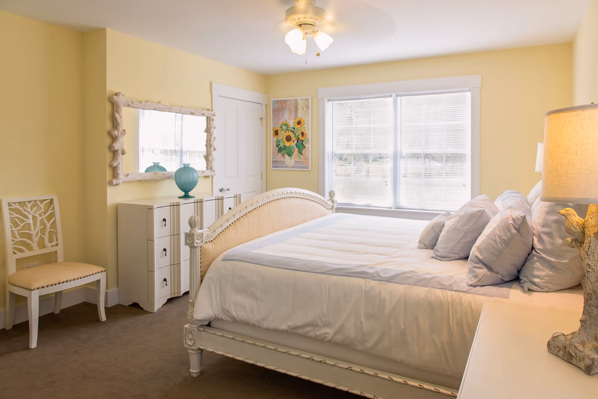 A bright bedroom with pale yellow walls featuring a large bed with white and light blue bedding and multiple pillows. There is a white dresser with a decorative blue vase and a large mirror above it. A white chair with a coral design is positioned against the wall. A window with blinds lets in natural light, and a sunflower painting hangs on the wall next to the window. A lamp with a textured base is on a bedside table.