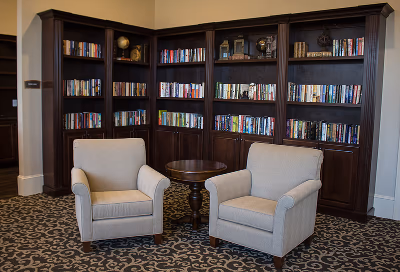 Two beige armchairs flank a small round wooden table in front of tall dark wood bookshelves filled with books.
