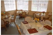 A dining room with several tables covered with white tablecloths, each set with plates, napkins, glasses, and silverware. The room has large windows with blinds allowing natural light to fill the space. Chairs with cushioned seats surround the tables, and there are small floral centerpieces on some tables.