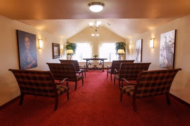 A small, cozy room with red carpet and six plaid upholstered chairs arranged in two rows facing a table with two lamps and plants near a window. The walls are decorated with framed artwork and wall sconces provide lighting.