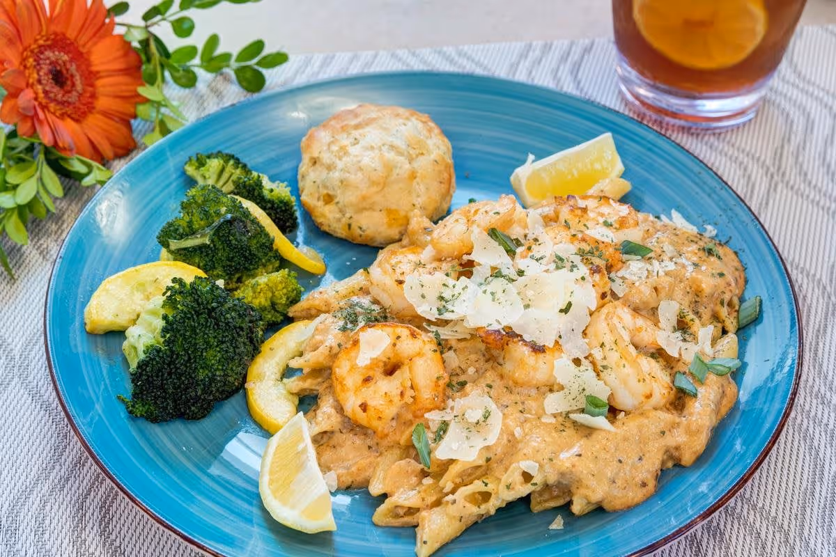 A blue plate with a serving of shrimp and creamy pasta topped with shaved cheese and herbs, steamed broccoli with lemon wedges, and a biscuit. A glass of iced tea with lemon is visible in the background, along with an orange flower and green leaves on the table.