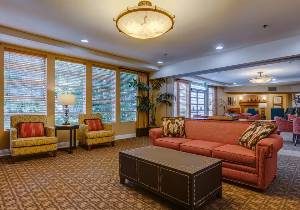 A spacious and well-lit living room area in a senior living facility with a red sofa adorned with patterned pillows, two yellow armchairs with red cushions, a dark rectangular coffee table, large windows with wooden blinds, a floor lamp, and potted plants. In the background, there is a dining area with multiple chairs and tables, a fireplace, and decorative wall art.