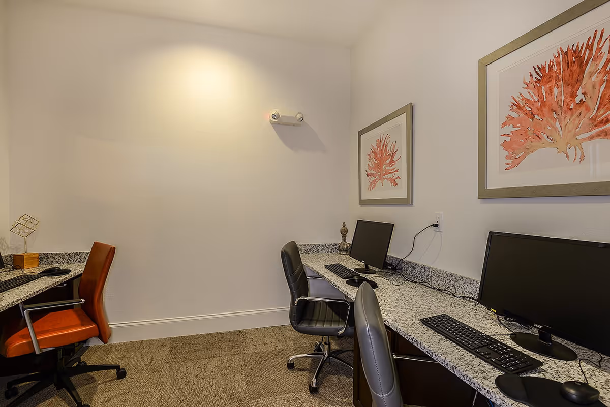 A small office or computer room with a granite countertop desk along two walls. There are three office chairs, two black and one orange, positioned in front of three computer monitors with keyboards and mice. The walls are white and decorated with framed artwork featuring red coral designs. The floor is carpeted in a neutral tone.