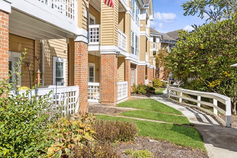 Exterior view of a senior living facility with a paved walkway, green grass, shrubs, and trees. The building features brick columns and beige siding with white trim and balconies.