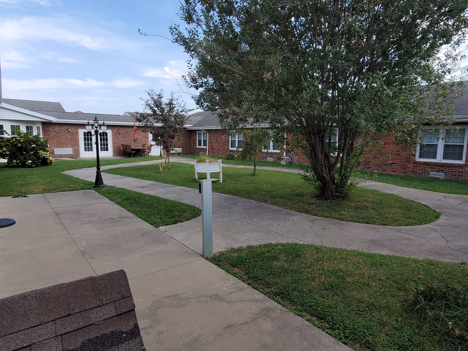 Brick single-story senior living buildings around a grassy courtyard with walkways, trees, a lamp post, and patio seating.