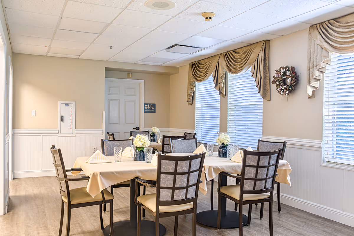 A dining room in a senior living facility with two tables covered with beige tablecloths, each set with glassware, folded napkins, and small flower arrangements in vases. The room has beige walls, large windows with blinds and decorative valances, and a wreath hanging on the wall. A door with a restroom sign is visible in the background.