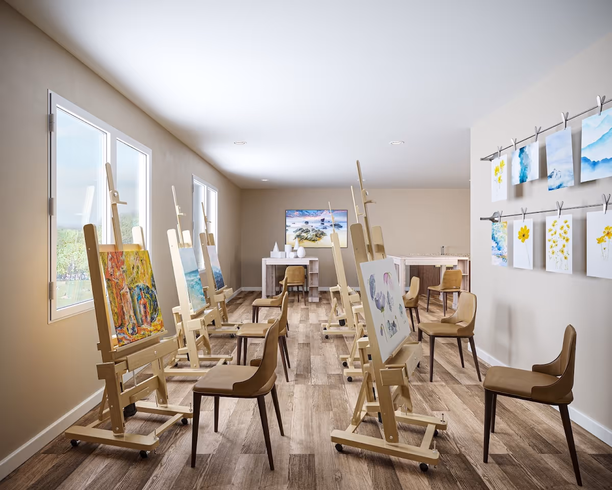 A bright art studio room with wooden easels holding paintings and brown chairs arranged in front of each easel. The room has large windows on one side letting in natural light, wooden flooring, and art pieces displayed on the wall. A table with decorative items is visible at the far end of the room.