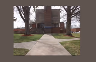 Front exterior view of a brick building with a central entrance, flanked by two large trees and a concrete walkway leading up to the door. A sign near the entrance reads 'Somerwoods Nursing and Rehabilitation Center'.