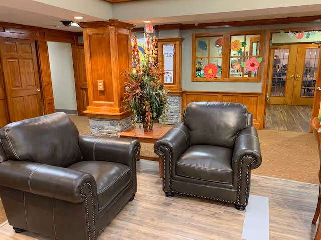 Two black leather armchairs positioned on a wooden floor in a cozy interior space with wood-paneled walls and a decorative floral arrangement on a small table between the chairs. The background features wooden doors, windows with colorful paper flower decorations, and a carpeted area.