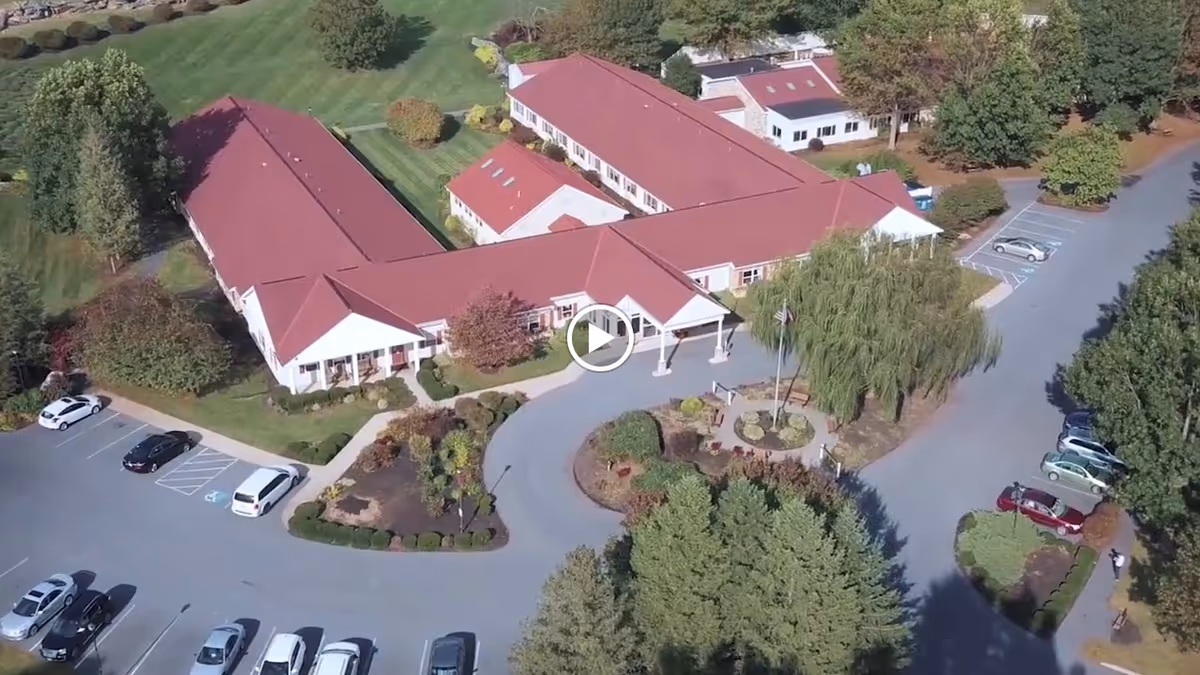 Aerial view of Oak Leaf Manor South senior living facility showing a large building with red roofs surrounded by landscaped gardens, trees, and parking lots with several parked cars.