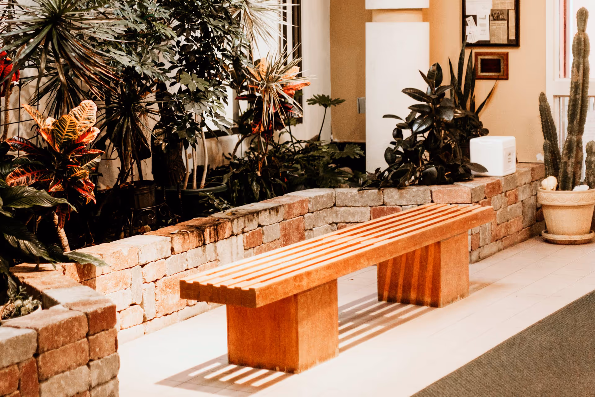 A wooden slatted bench in an indoor plant-filled seating area with brick planters and potted plants.