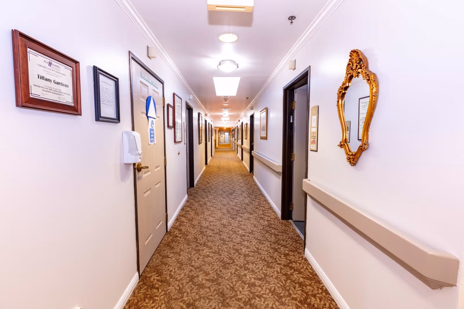 A long, well-lit hallway in a senior living facility with beige patterned carpet, white walls, and several closed doors on both sides. The walls are decorated with framed certificates, notices, and a gold ornate mirror. Handrails run along both sides of the hallway.