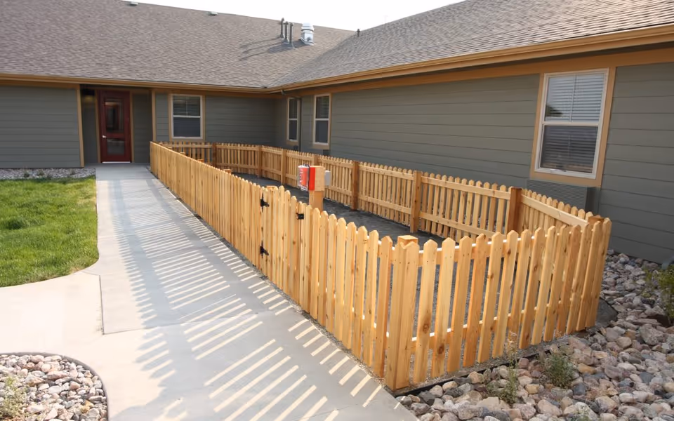 Outdoor area of Mountain Plaza Assisted Living & Memory Care featuring a wooden fenced garden or patio space adjacent to a building with green siding and a brown roof. There is a concrete walkway leading to a door, with rocks and grass surrounding the fenced area.