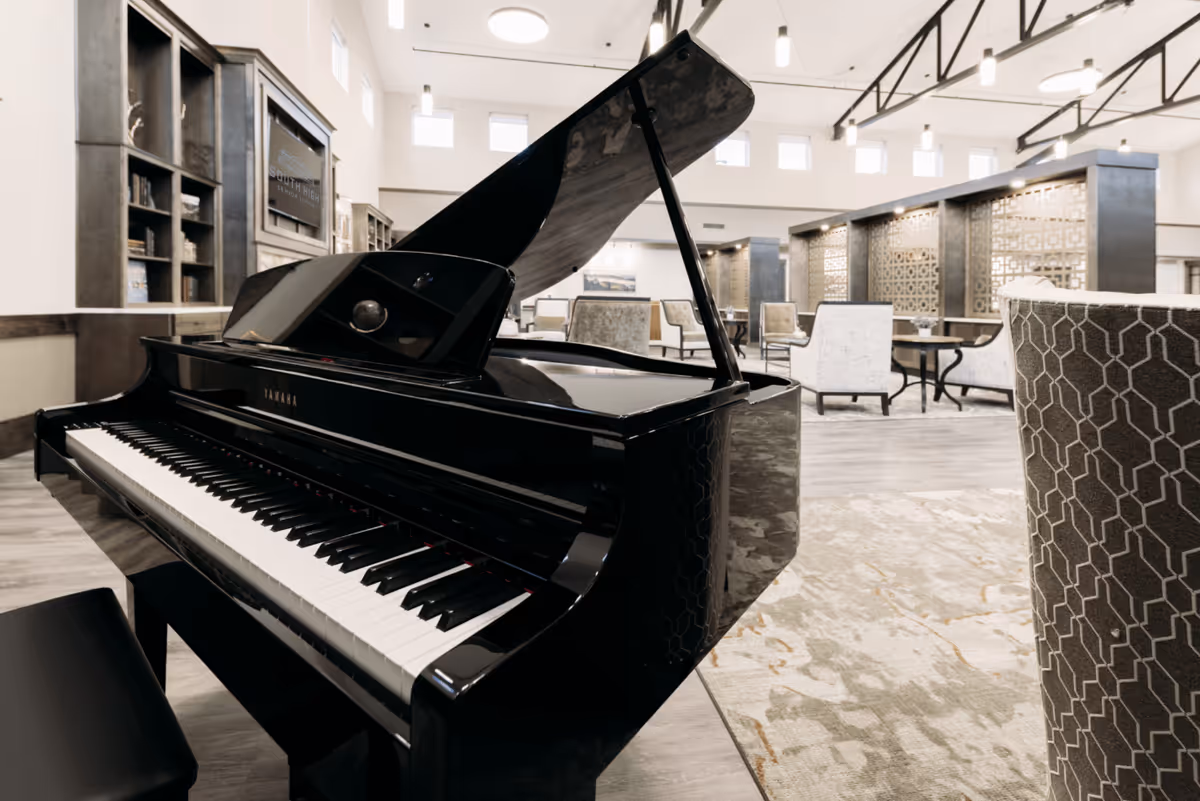 A glossy black grand piano sits in the foreground of a spacious senior living lounge with chairs, tables, and high ceilings.