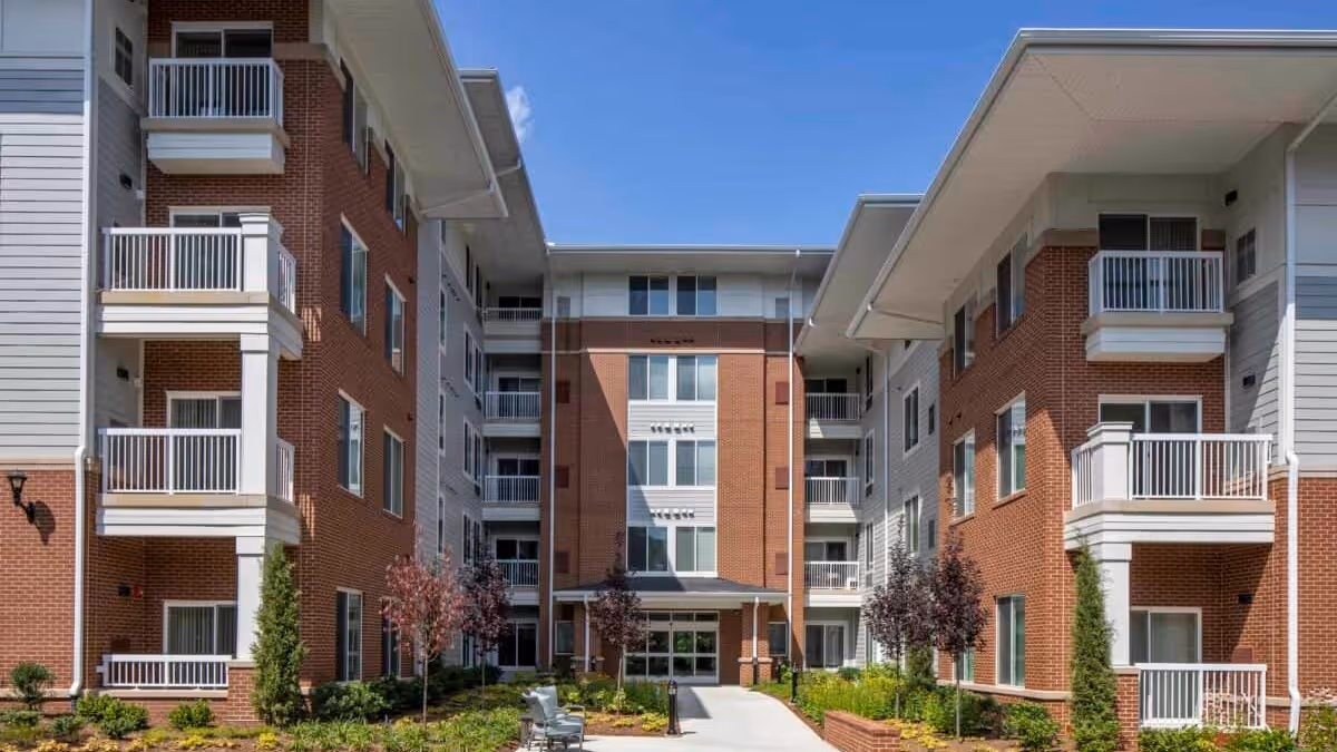 Exterior view of a multi-story senior living facility building with red brick and white siding. The building has balconies with white railings and a central entrance pathway lined with small trees and shrubs under a clear blue sky.