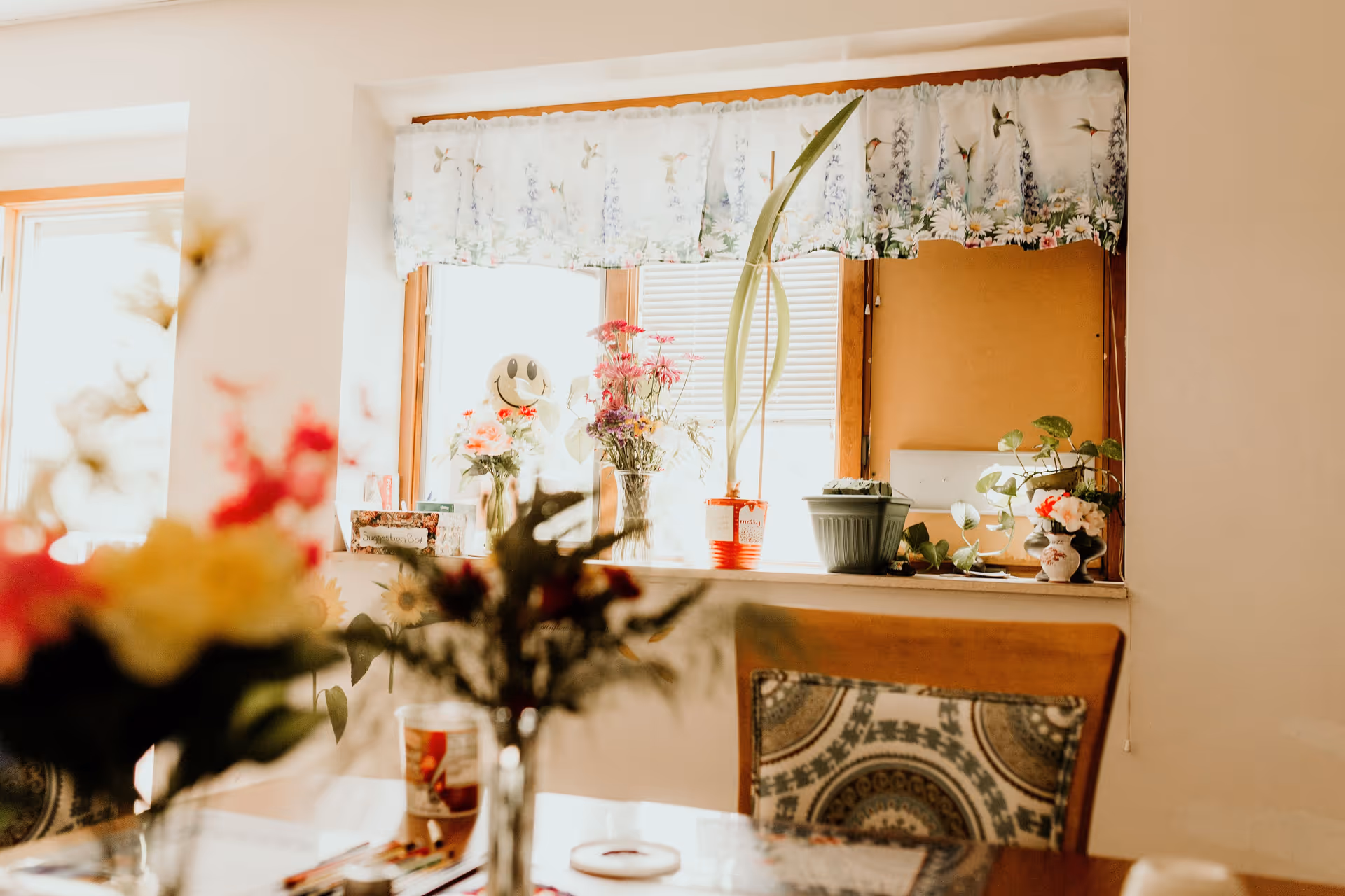 A bright dining area with a wooden table and chairs. The window sill is decorated with various potted plants and flowers, and a window with floral curtains lets in natural light.
